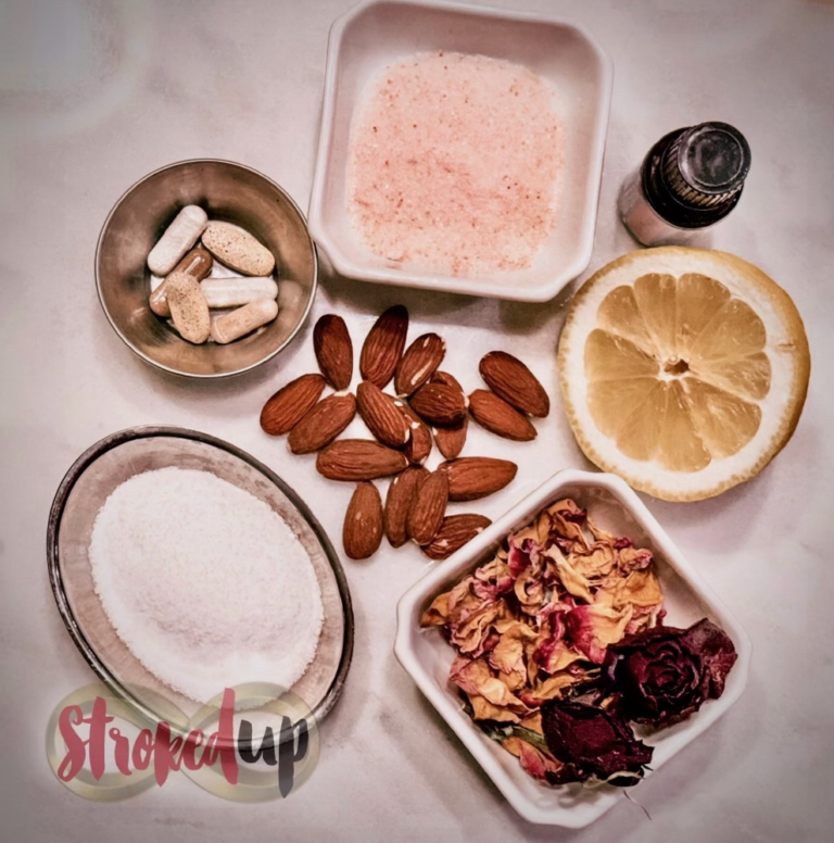 against a white marble backdrop, small bowls of sea salt, pink salt, rose petals, and a half a lemon frame a handful of almonds next to a bottle of wild orange essential oil.